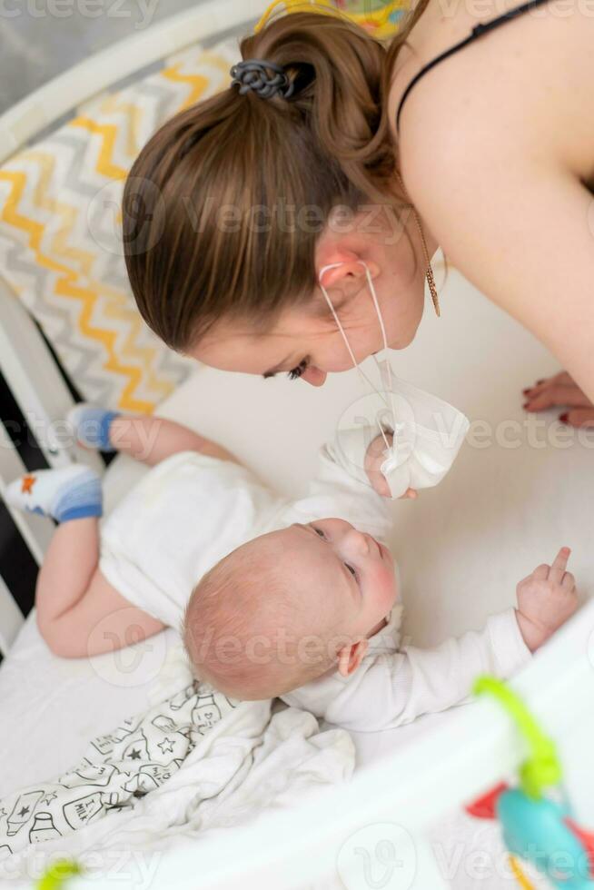 A mother in a medical protective mask takes care of her baby at home in a corontine. Prophylactic protection of coronavirus and covid-19 during a pandemic. photo