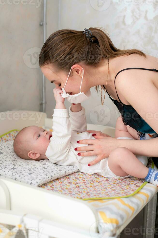 A mother in a medical protective mask takes care of her baby at home in a corontine. Prophylactic protection of coronavirus and covid-19 during a pandemic. photo