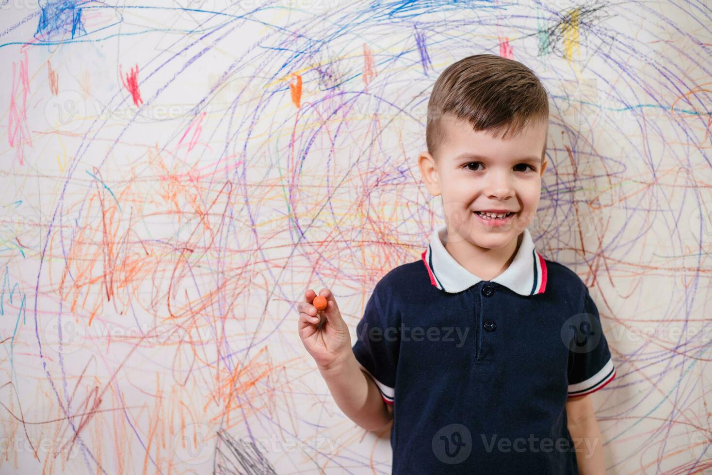 A cheerful boy with a pencil stands near the wall that he painted. A child is engaged in creativity at home. photo