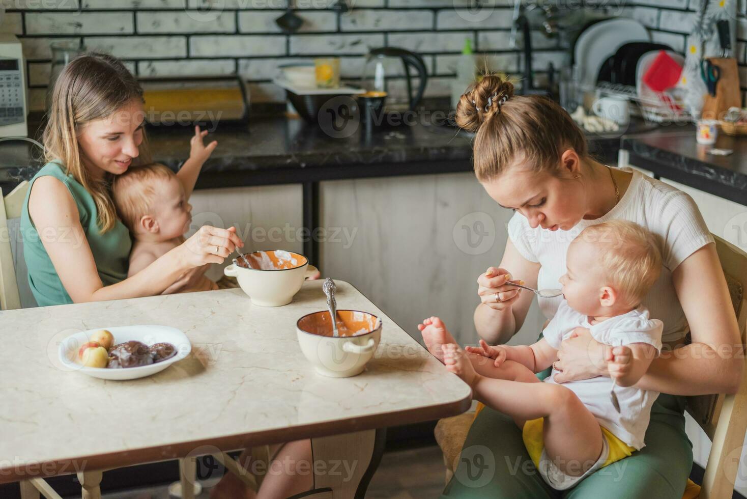 Two tired but happy mothers feed their babies milk porridge in the kitchen photo