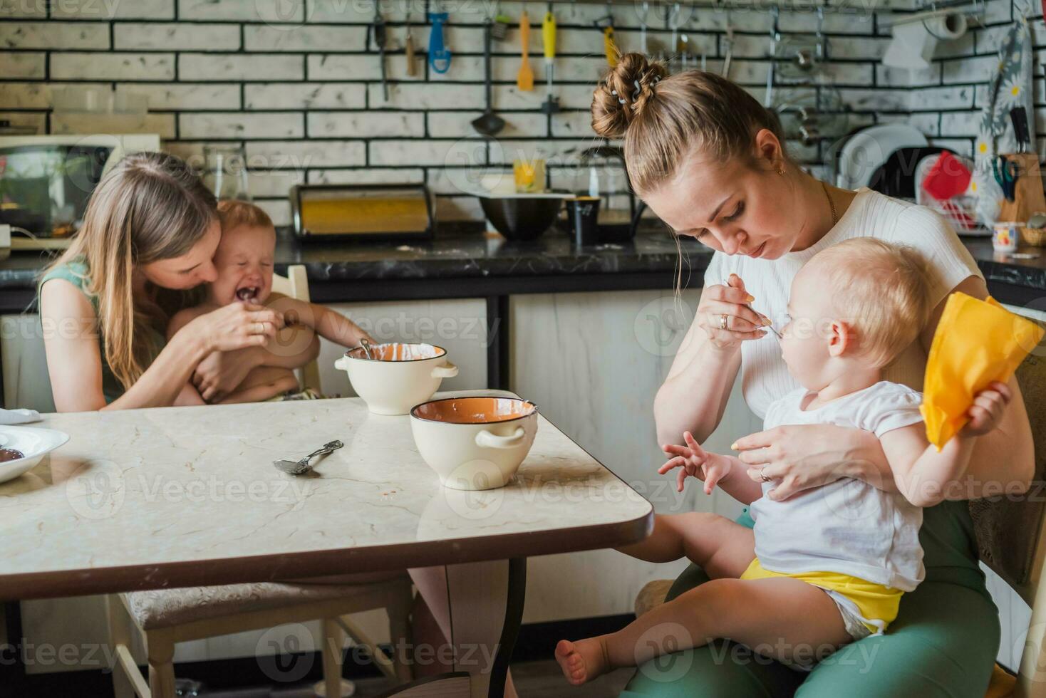 Two young mothers together feed their happy babies milk porridge in the kitchen photo