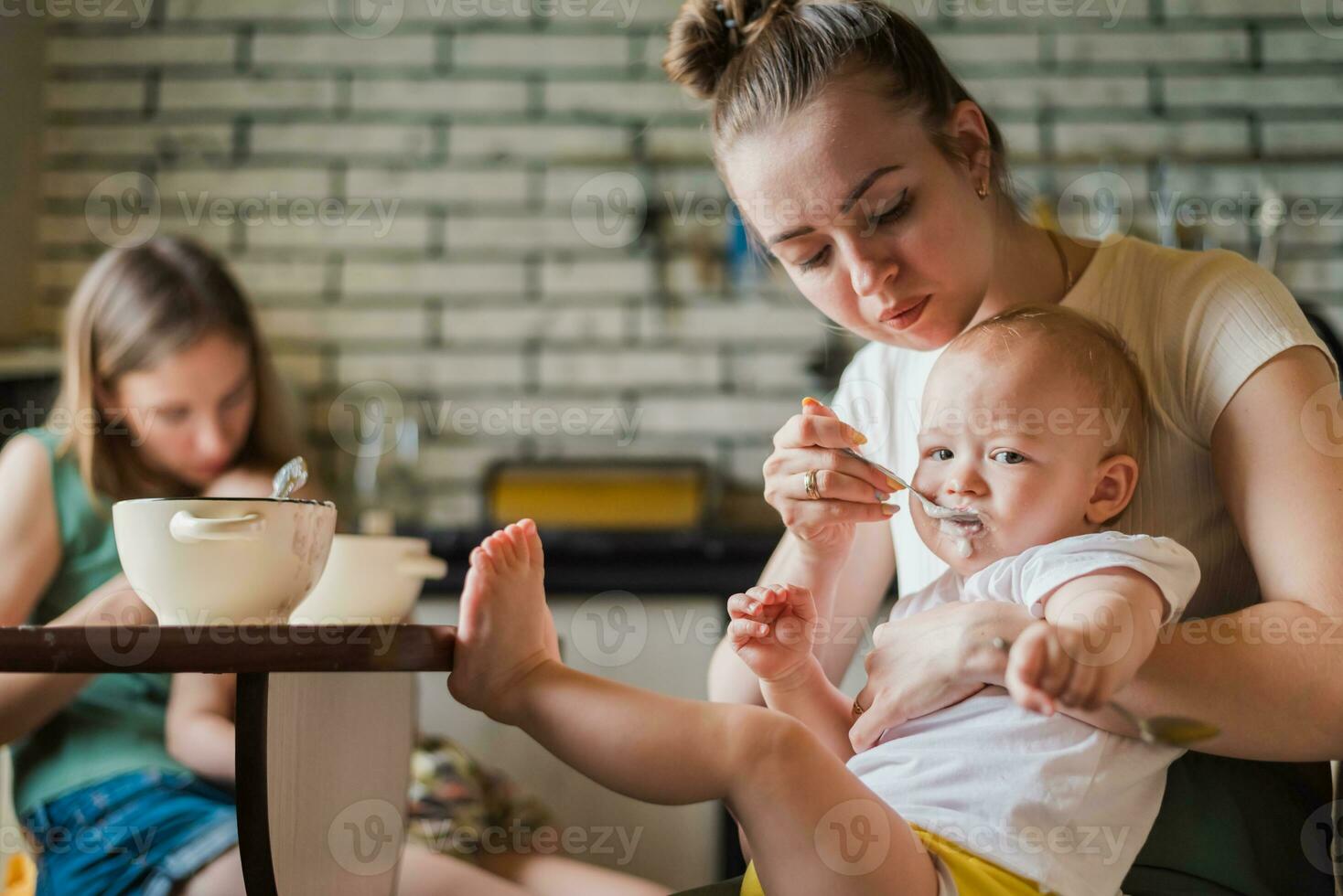 Two tired but happy mothers feed their babies milk porridge in the kitchen photo