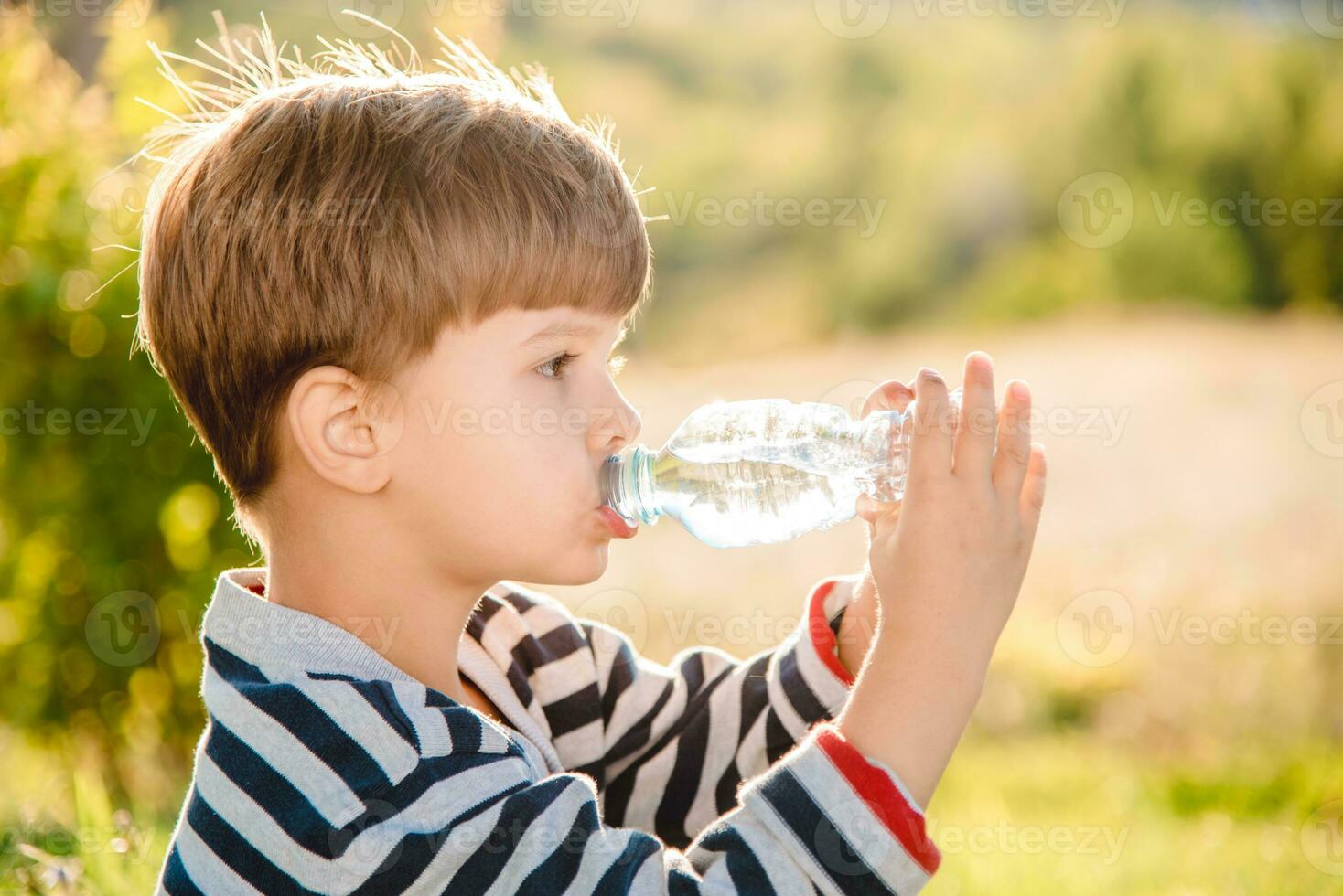 A beautiful child sitting on the grass drinks water from a bottle in the summer at sunset. Boy quenches his thirst on a hot day photo