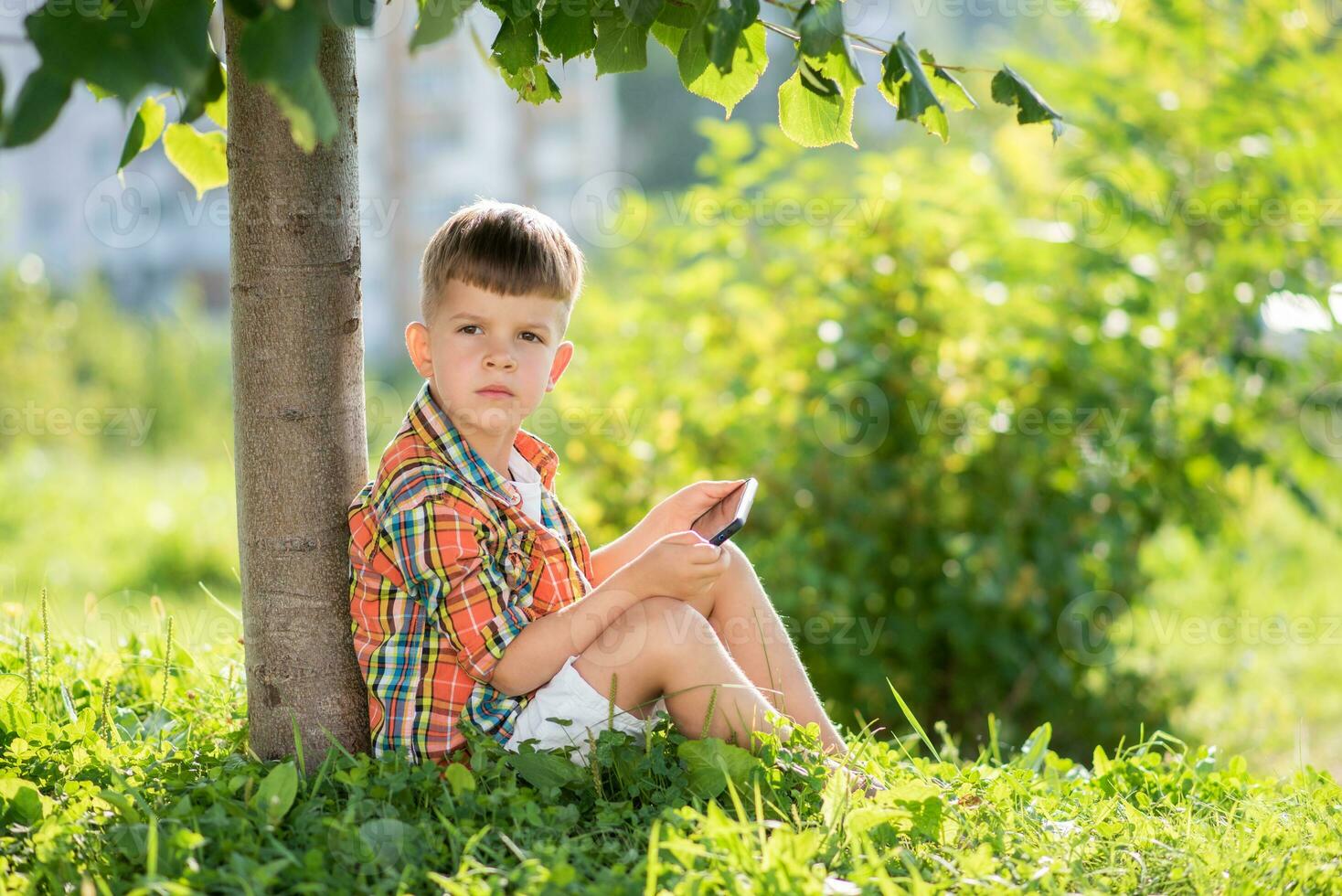 A beautiful child sits on the grass and looks at the phone in the summer at sunset. Cute boy with mobile in hand photo