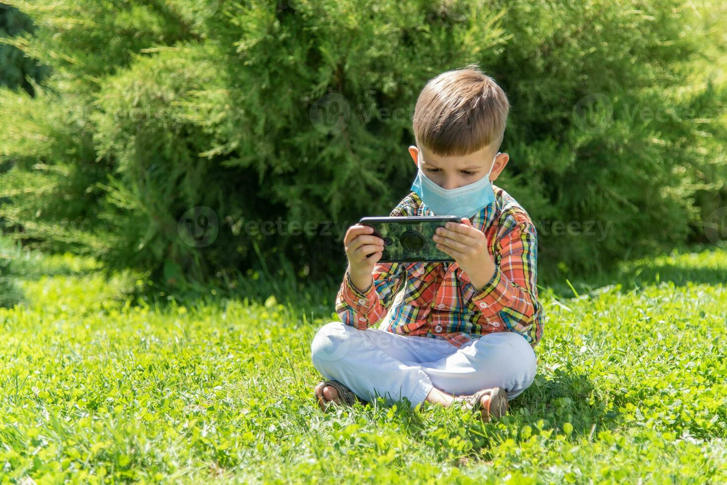 A kid in a medical mask sits on the grass and looks in the phone cartoons in the summer at sunset. Child with a mobile phone in his hands. Prevention against coronavirus Covid-19 during a pandemic photo