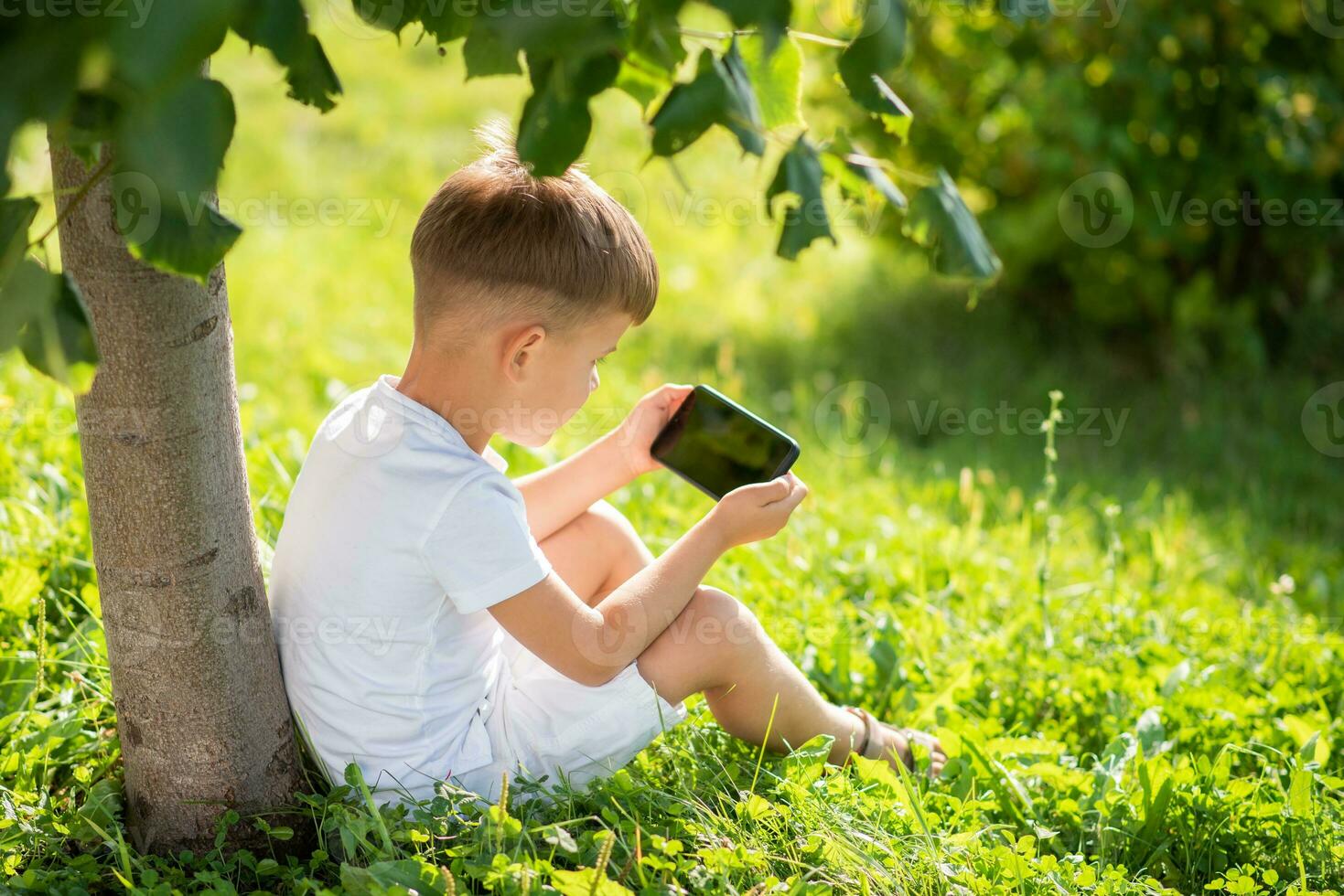 Cheerful boy sitting on the grass looks cartoons in the phone in the summer at sunset. Cute baby having fun in nature photo