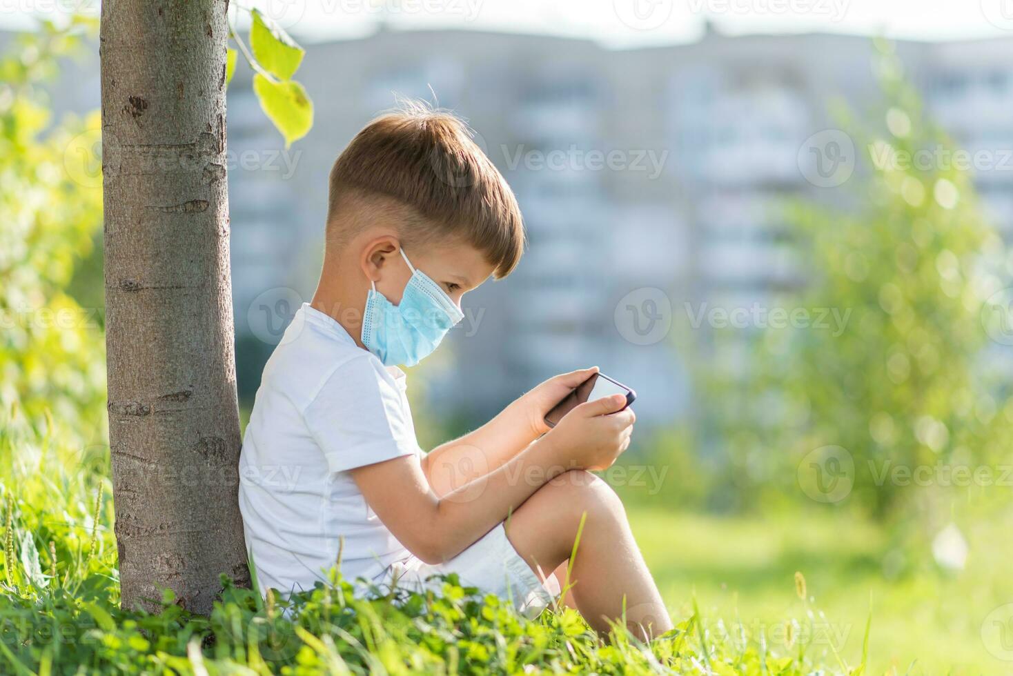 A kid in a medical mask sits on the grass and looks in the phone cartoons in the summer at sunset. Child with a mobile phone in his hands. Prevention against coronavirus Covid-19 during a pandemic photo