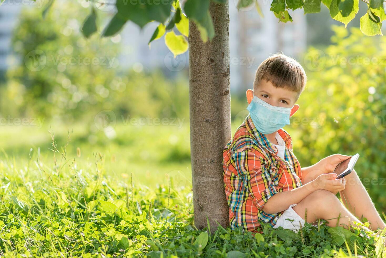 A kid in a medical mask sits on the grass and looks in the phone cartoons in the summer at sunset. Child with a mobile phone in his hands. Prevention against coronavirus Covid-19 during a pandemic photo