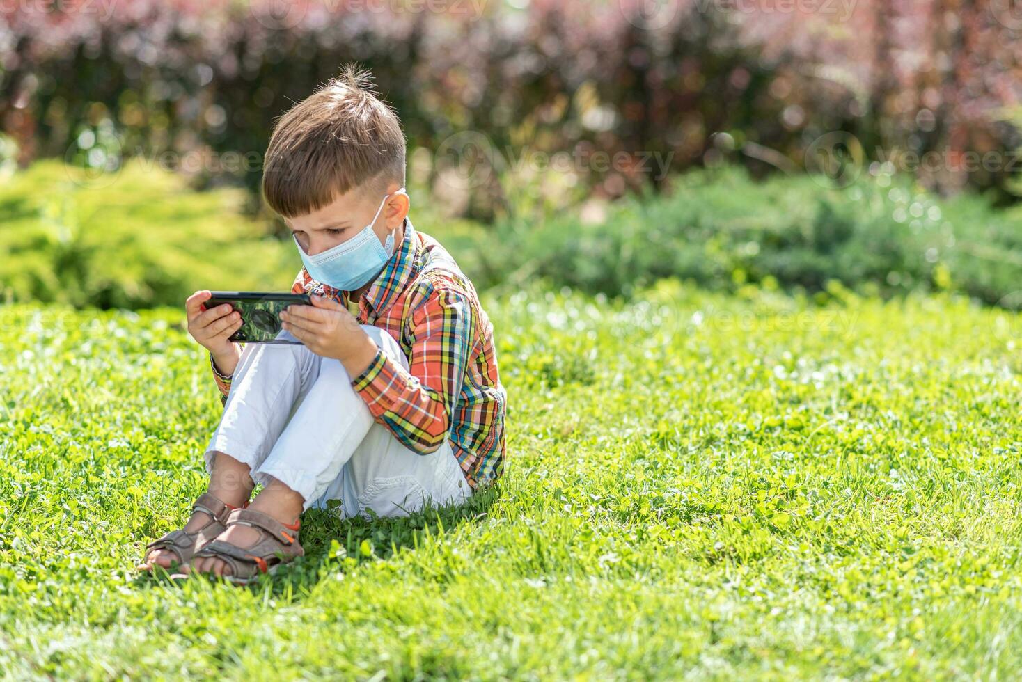 A kid in a medical mask sits on the grass and looks in the phone cartoons in the summer at sunset. Child with a mobile phone in his hands. Prevention against coronavirus Covid-19 during a pandemic photo