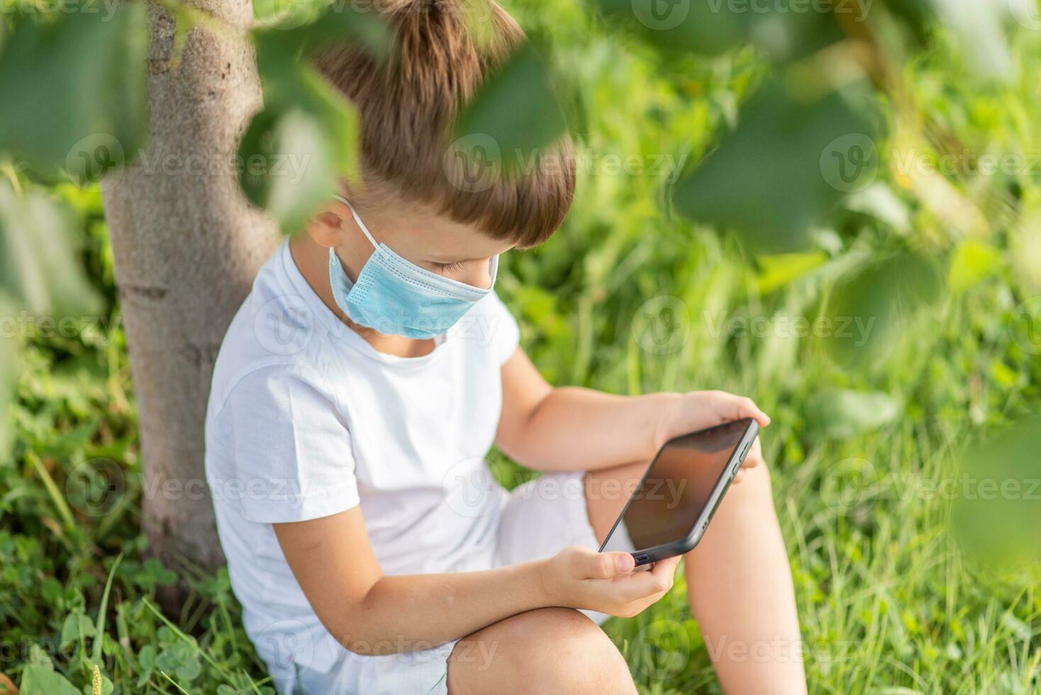 A kid in a medical mask sits on the grass and looks in the phone cartoons in the summer at sunset. Child with a mobile phone in his hands. Prevention against coronavirus Covid-19 during a pandemic photo