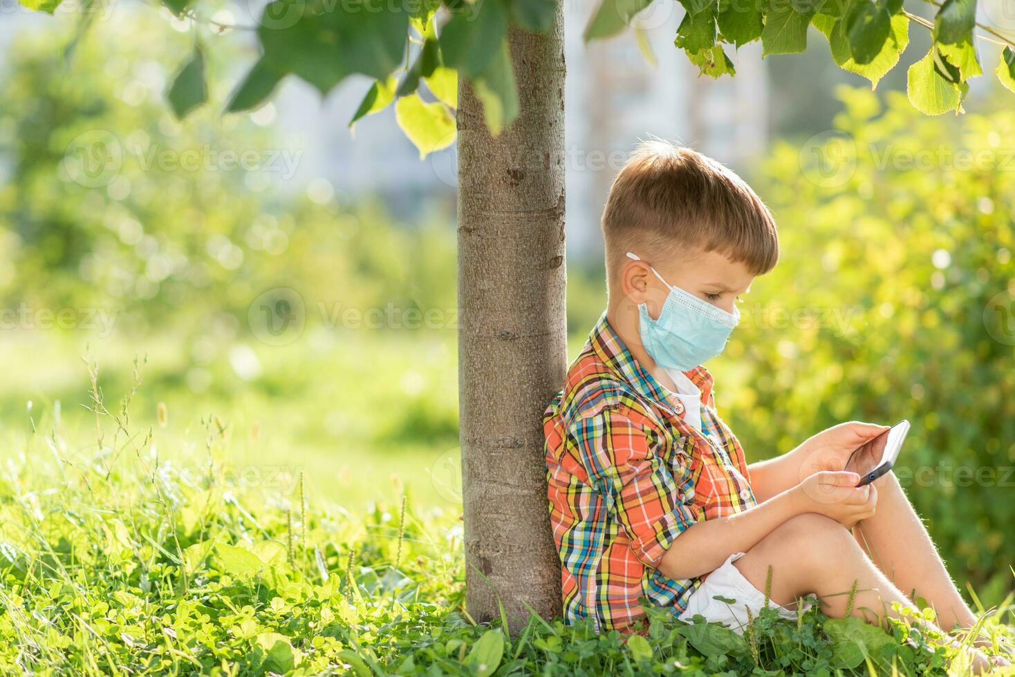 A kid in a medical mask sits on the grass and looks in the phone cartoons in the summer at sunset. Child with a mobile phone in his hands. Prevention against coronavirus Covid-19 during a pandemic photo