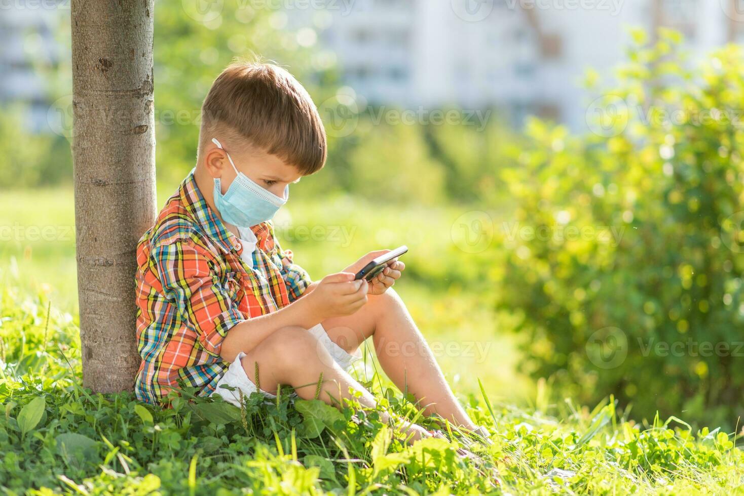 A kid in a medical mask sits on the grass and looks in the phone cartoons in the summer at sunset. Child with a mobile phone in his hands. Prevention against coronavirus Covid-19 during a pandemic photo