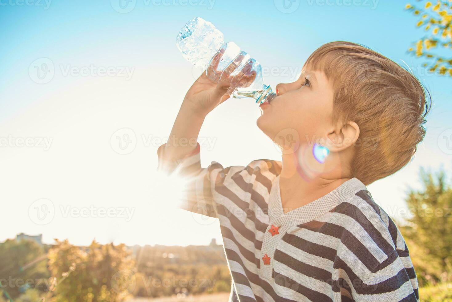 A beautiful child sitting on the grass drinks water from a bottle in the summer at sunset. Boy quenches his thirst on a hot day photo