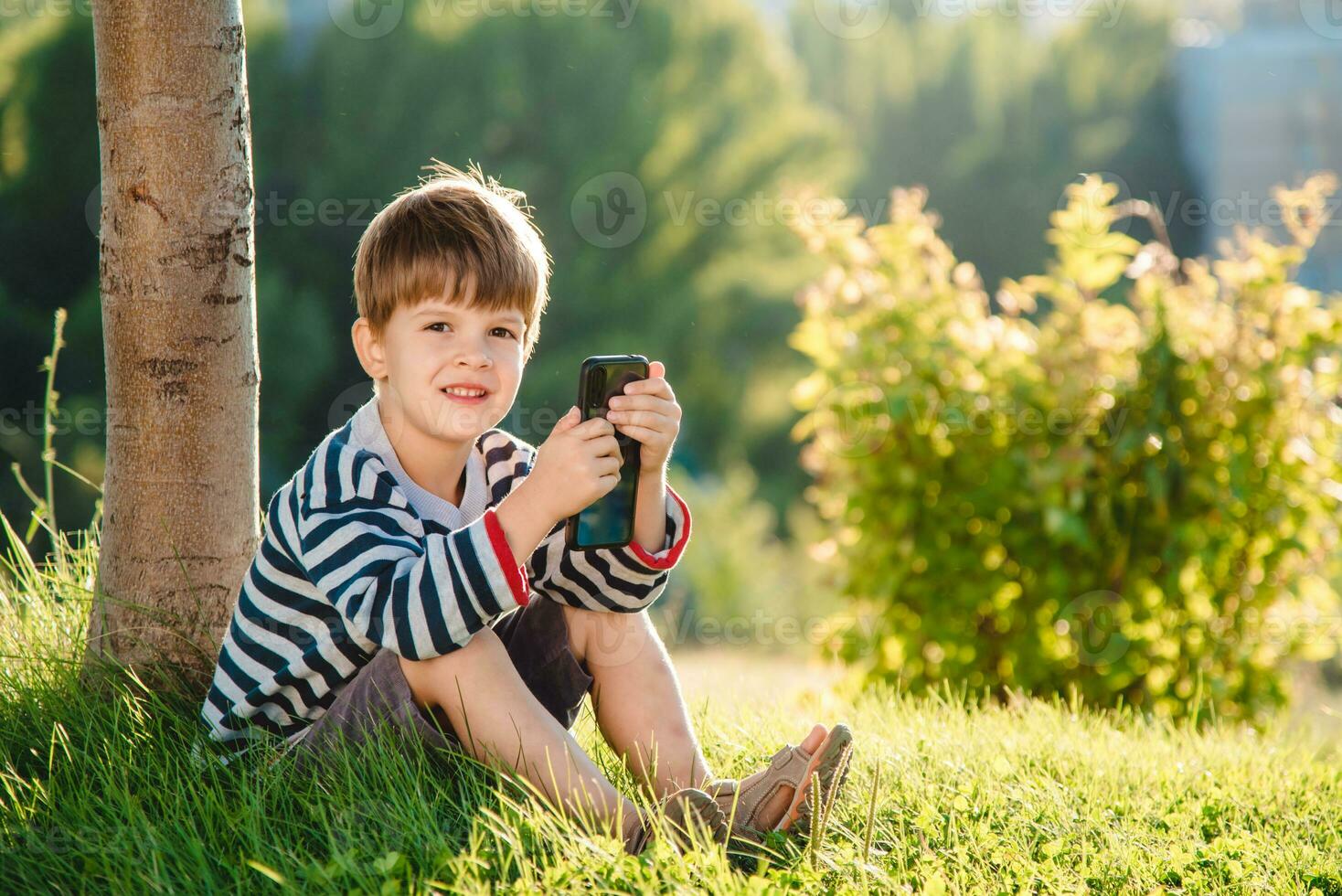 Cheerful boy sitting on the grass looks cartoons in the phone in the summer at sunset. Cute baby having fun in nature photo