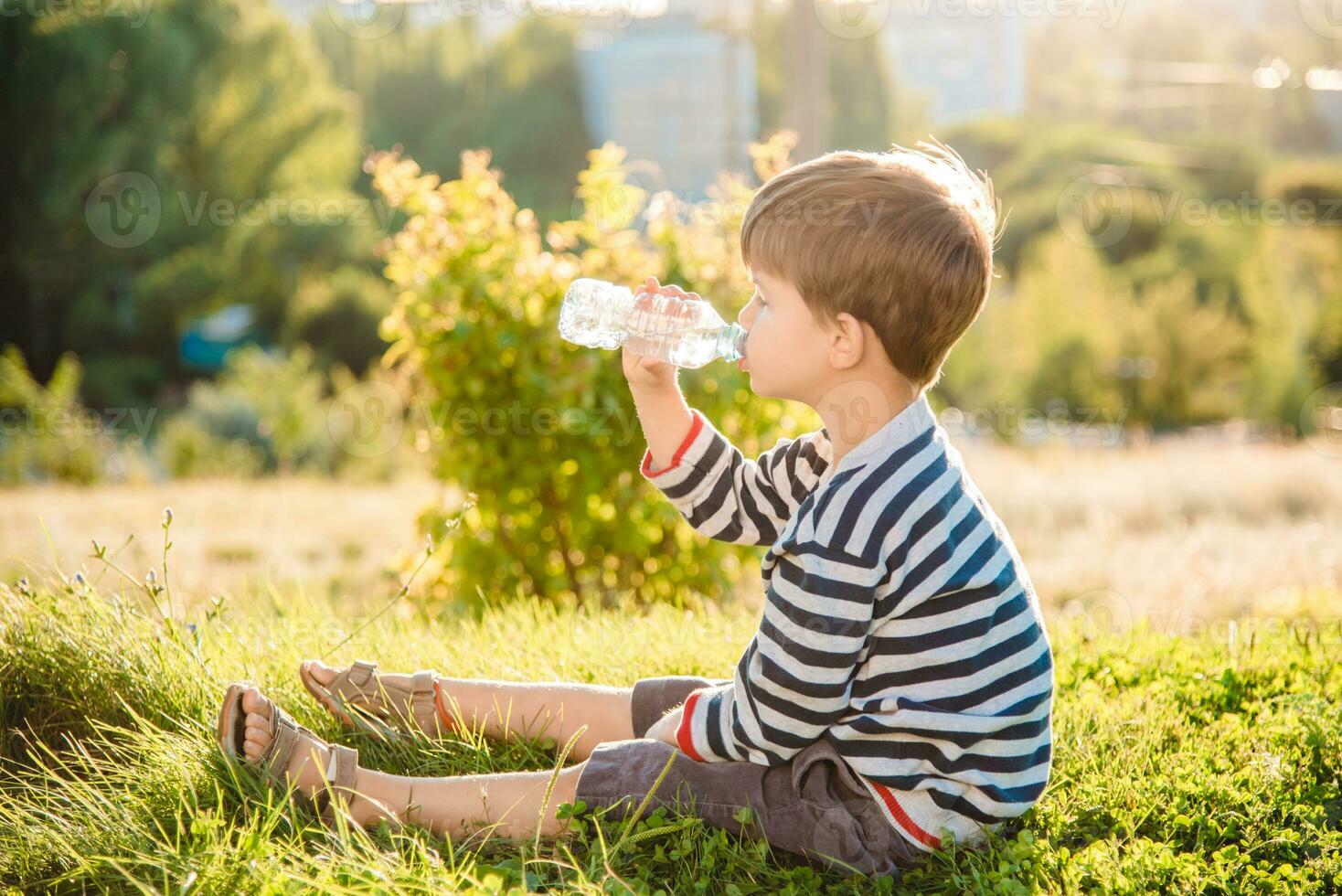 A beautiful child sitting on the grass drinks water from a bottle in the summer at sunset. Boy quenches his thirst on a hot day photo