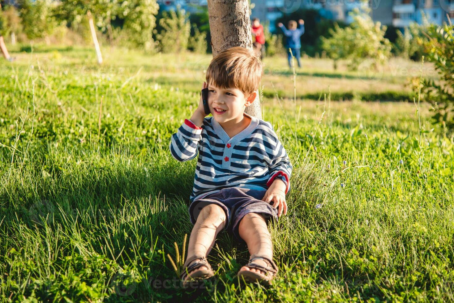 linda chico sentado en el césped habla por teléfono en el verano a puesta de sol. el niño comunica en un móvil foto