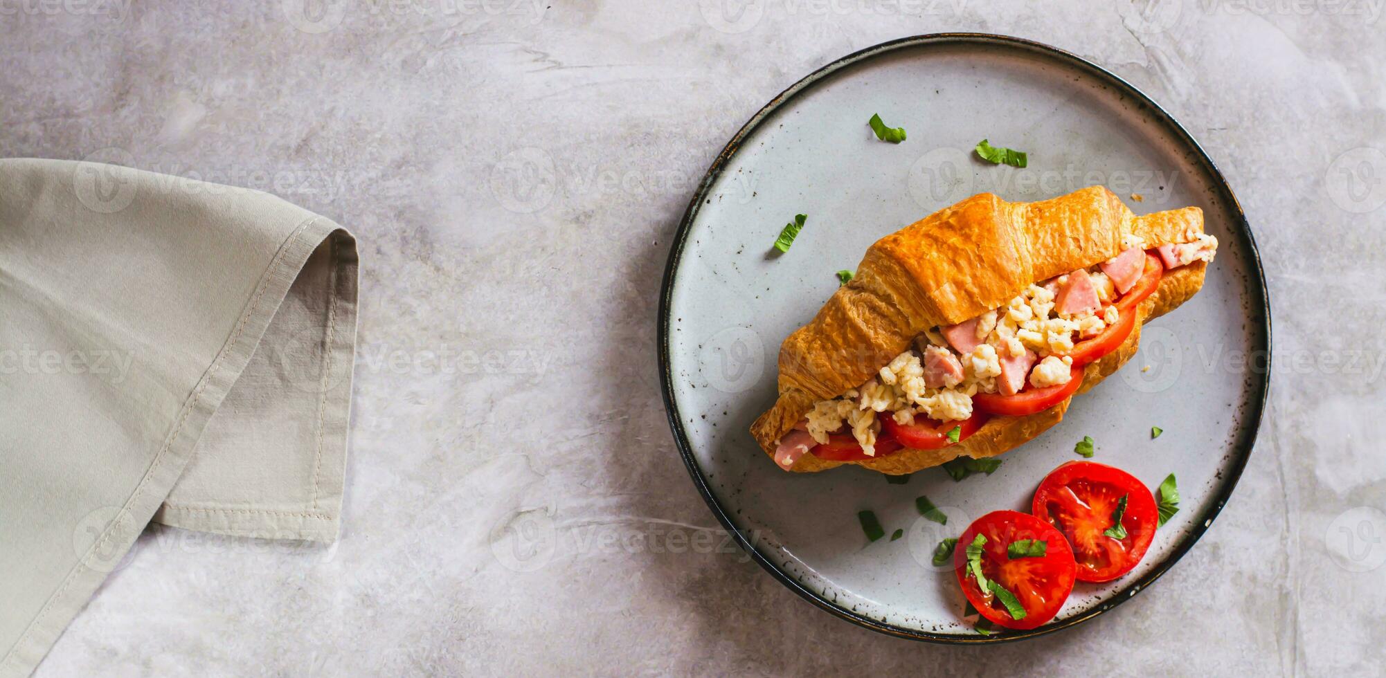 Croissant sandwich with scrambled eggs, sausages and tomatoes on a plate top view web banner photo