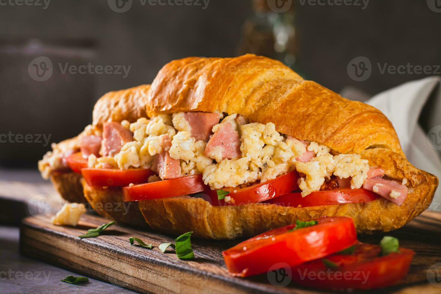 Close up of croissant sandwich with scrambled eggs, sausages and tomatoes on a board photo