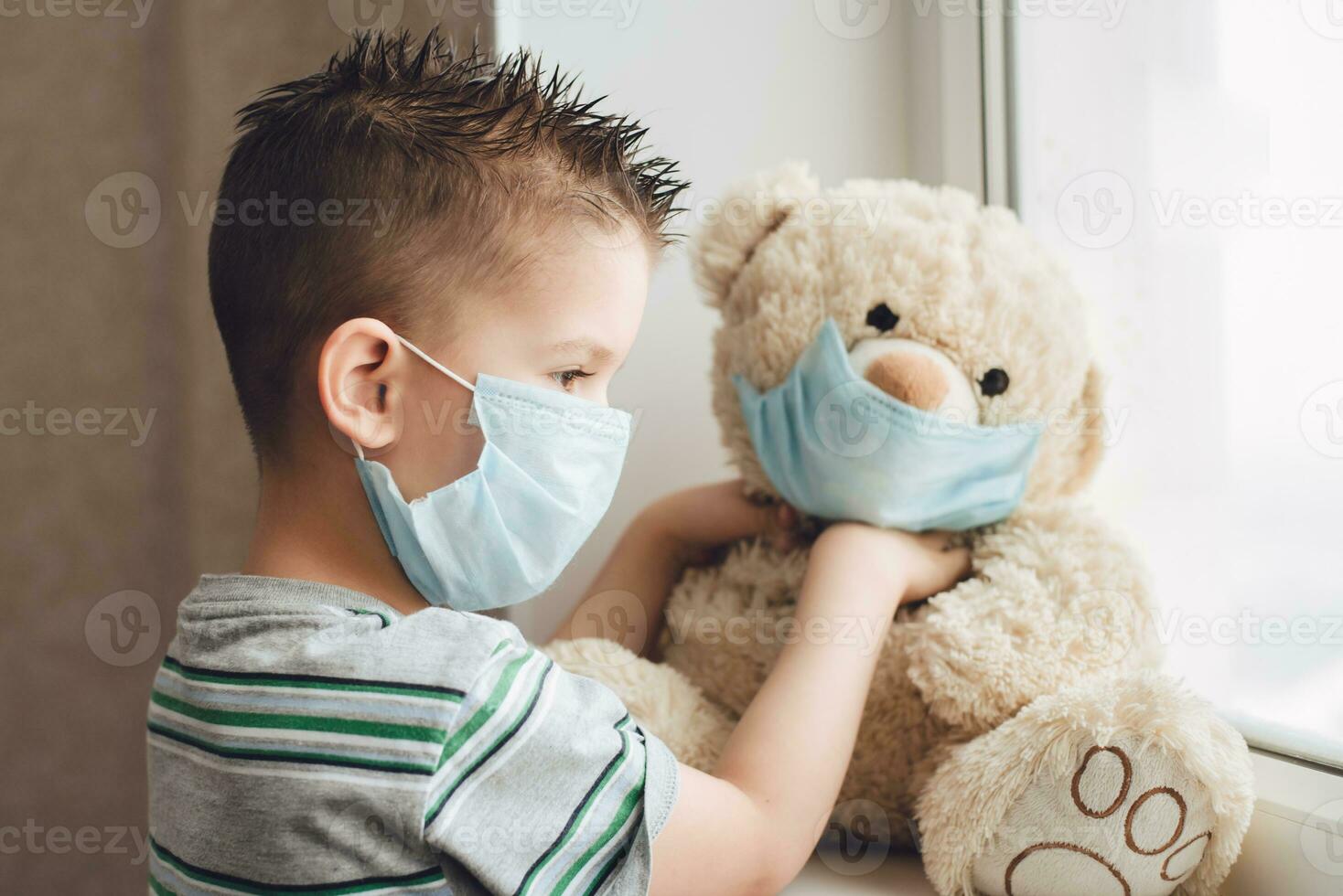 a small child in a mask sits at home in quarantine and looks out the window in a place with a teddy bear. Prevention of Coronavirus and Covid - 19. Concept photo