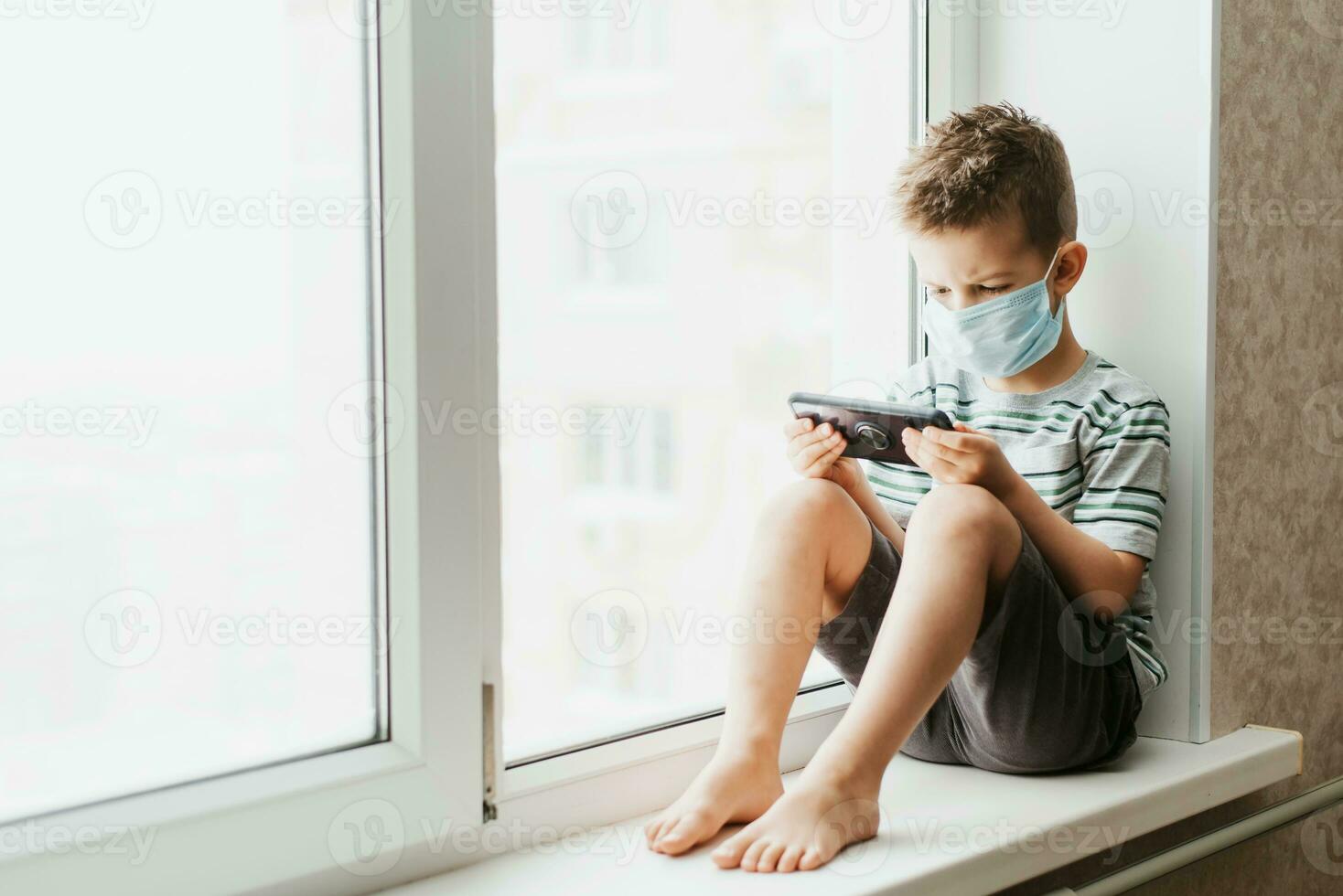 A cute child in a medical mask sits at home in quarantine by the window with a phone in his hands and looks into it. Prevention of coronavirus and Covid - 19 photo