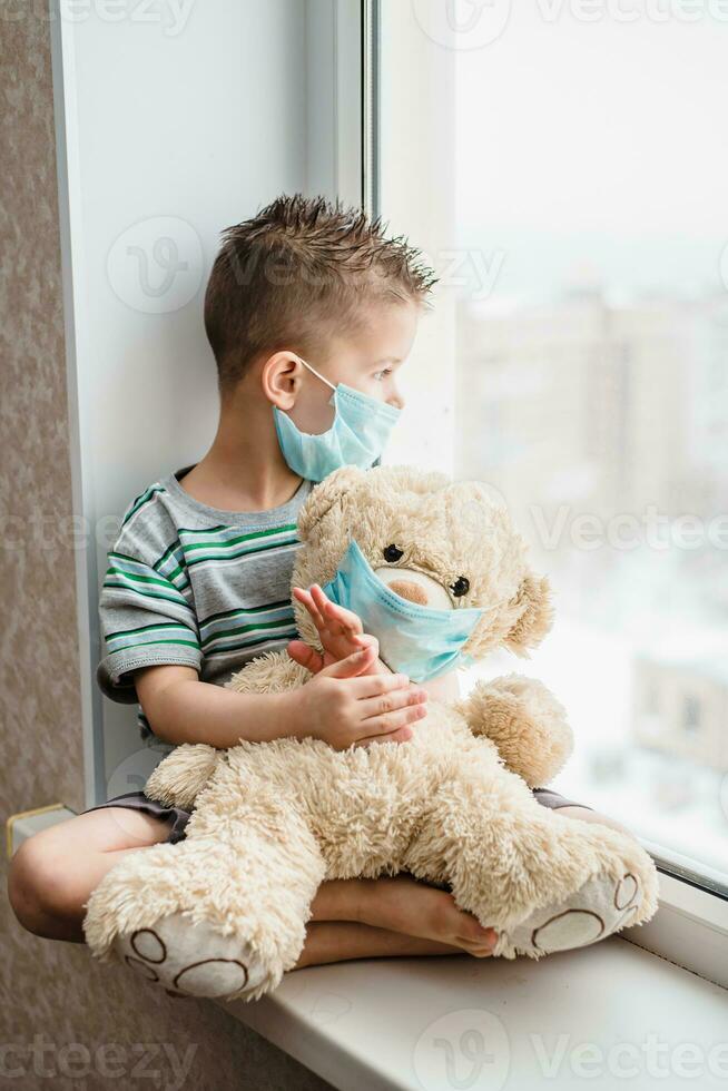 a small child in a mask sits at home in quarantine and looks out the window in a place with a teddy bear. Prevention of Coronavirus and Covid - 19. Concept photo