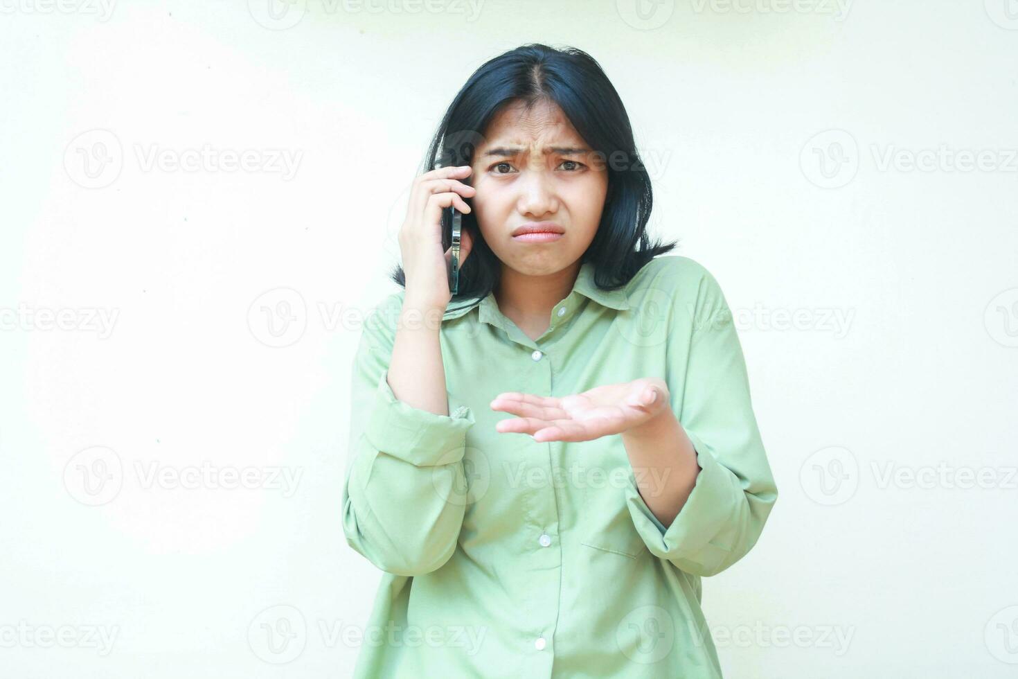 despistado asiático mujer vistiendo casual terminado tamaño verde camisa levantamiento palmas demostración yo hacer no saber gesto mientras Hablando en teléfono inteligente aislado en blanco fondo, qué hacer usted desear, mirando a cámara foto