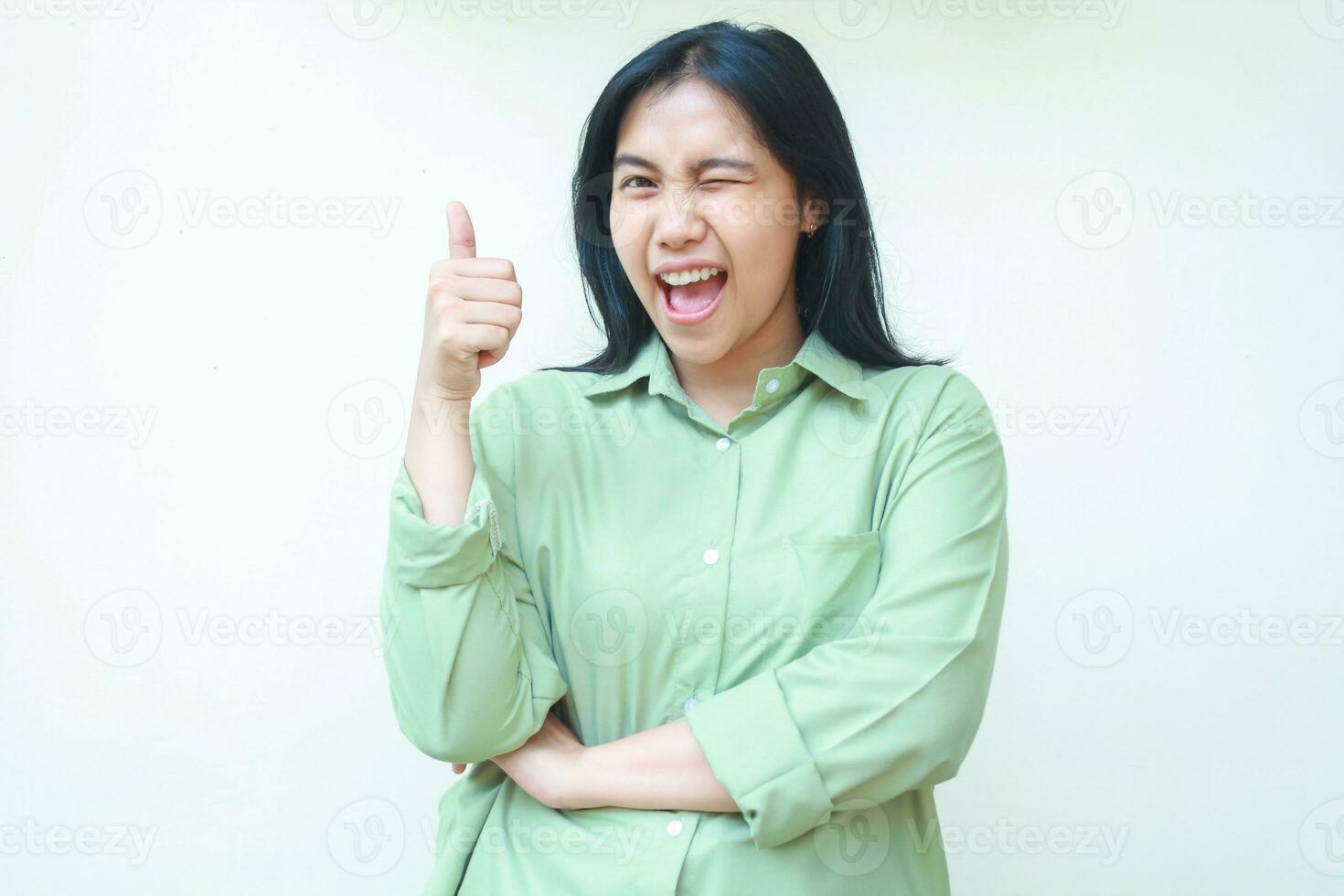playful optimistic asian woman with dark hair showing thumbs up shouting satisfied flirting to camera with eye closed wearing green over size clothes isolated on white background photo