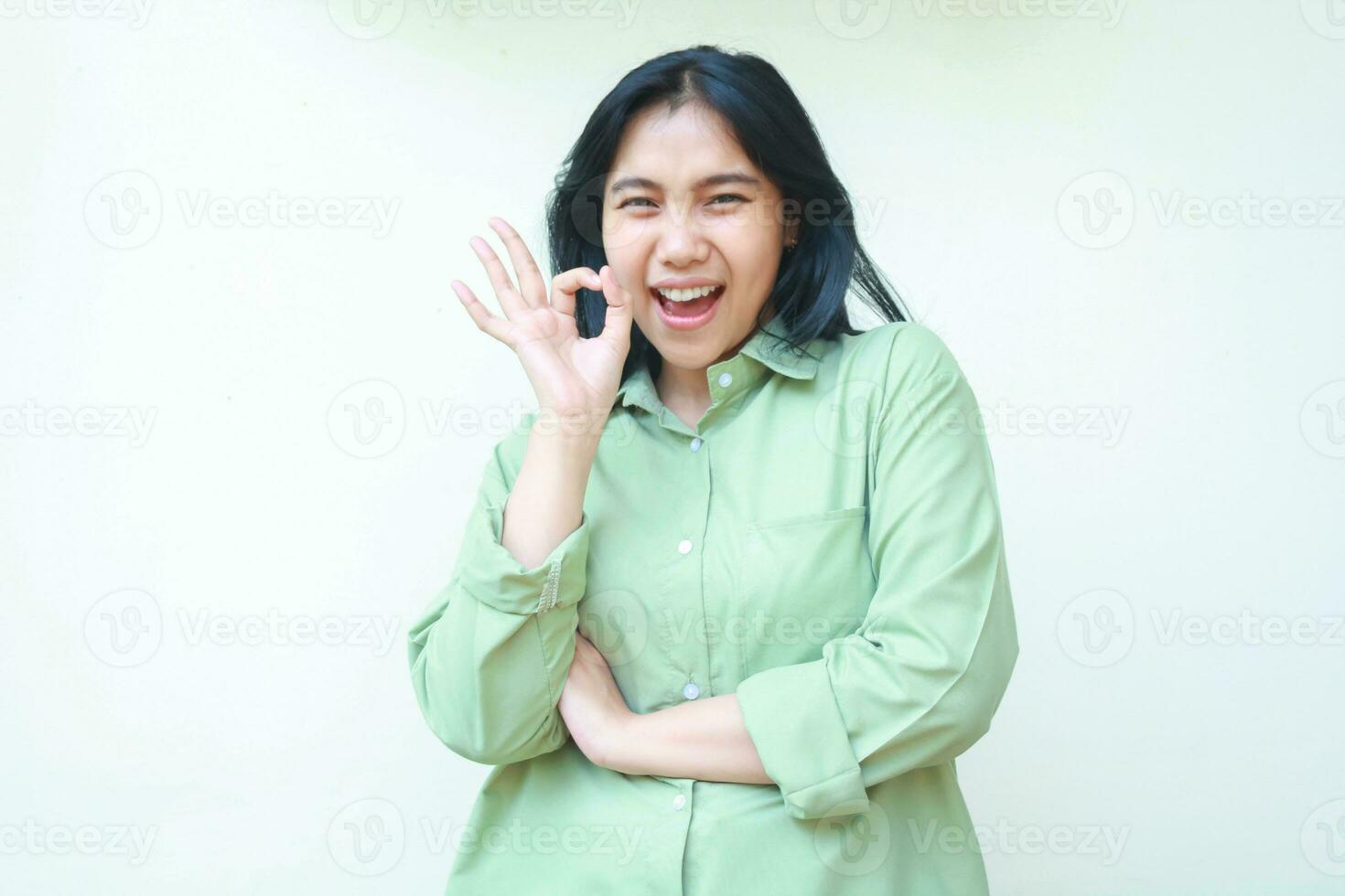 Encantado asiático mujer con oscuro pelo vistiendo verde terminado Talla ropa demostración Okay señales con plegable brazos mirando a cámara gritando satisfecho aislado en blanco antecedentes foto
