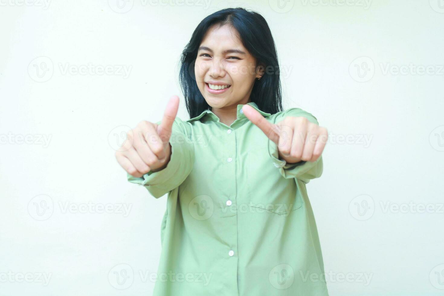 pleased dark hair asian woman showing thumbs up at camera with smiling satisfied face expression wearing green over size clothes standing over isolated white background photo