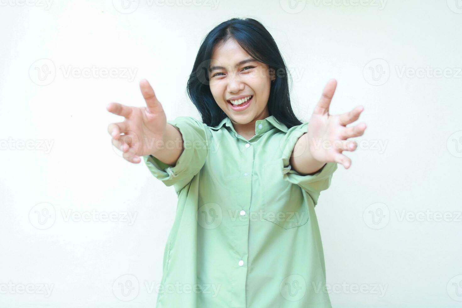 playful excited asian woman with dark hair spread hands giving hug gesture wearing green oversized shirt, welcoming you, looking at camera standing isolated white background photo