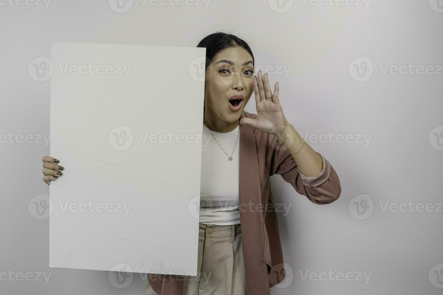 Young beautiful Asian woman employee wearing cardigan is shouting and screaming loud with a hand on her mouth while holding empty blank board, isolated by white background. photo