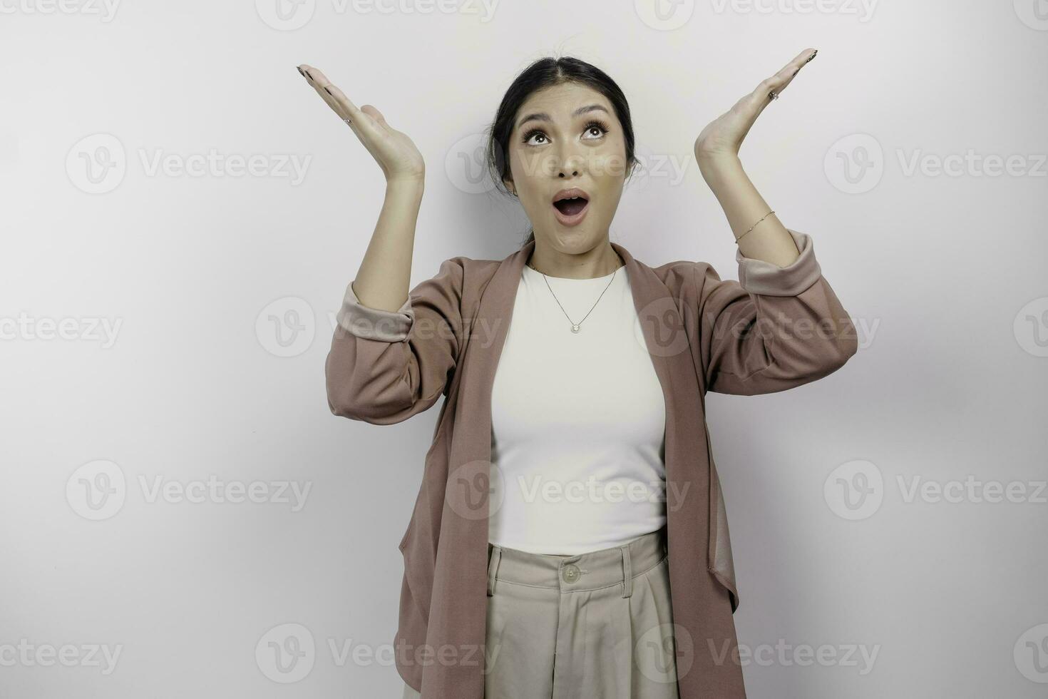Shocked Asian woman employee wearing cardigan pointing at the copy space on top of her, isolated by white background photo