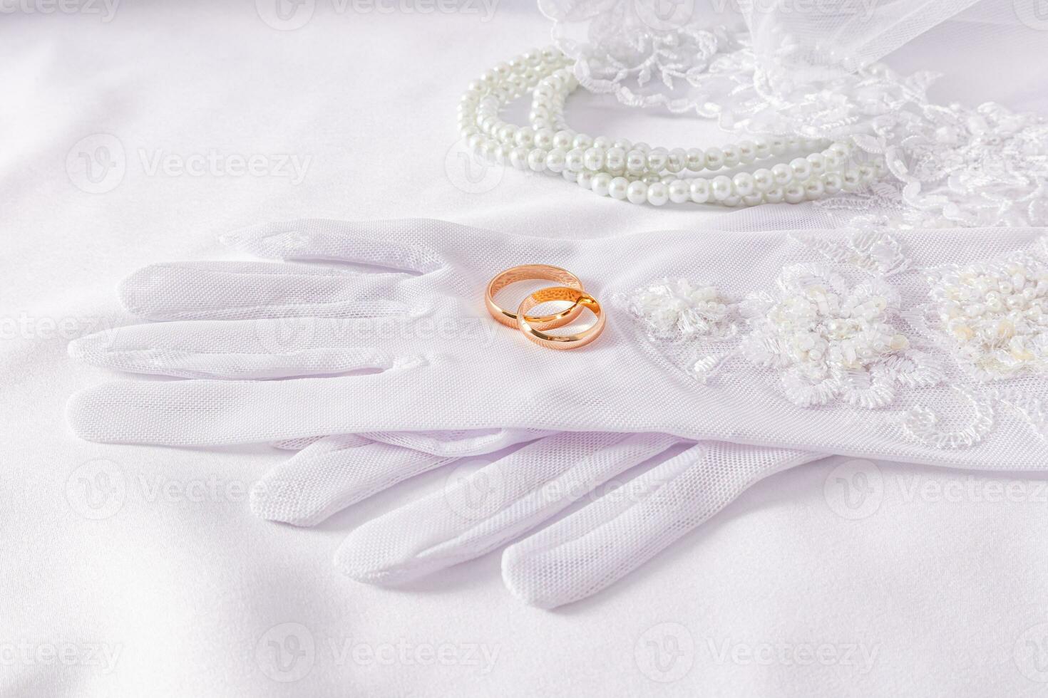 A pair of gold wedding rings lie on luxurious wedding white gloves. White satin background with pearl beads. Wedding accessories concept. photo