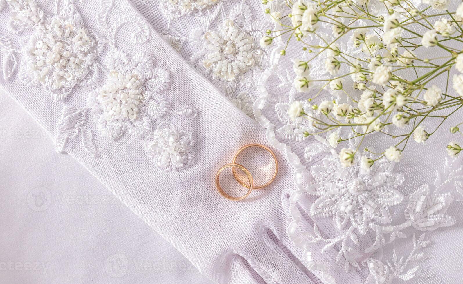 Two gold wedding rings lie on the bride's chic beaded gloves embroidered with beads and sequins. Top view. Satin white background. photo