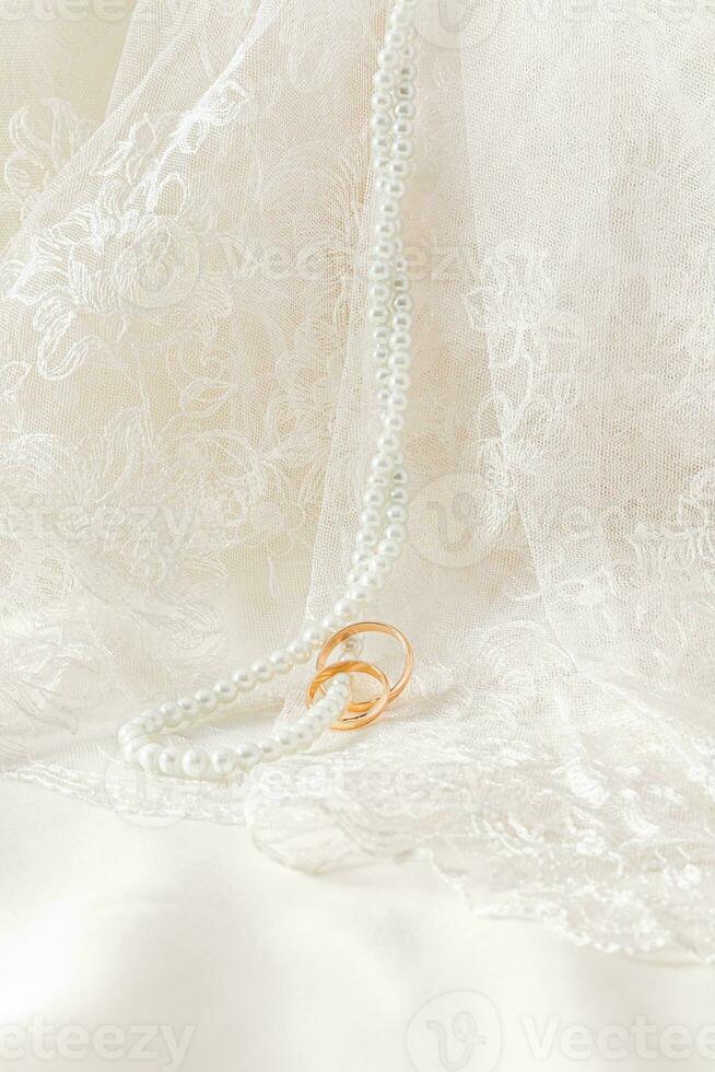 Retro beige tulle and long pearl beads hang along the soft folds of fabric with gold wedding rings worn on them. Vertical view. A copy space. mockup photo