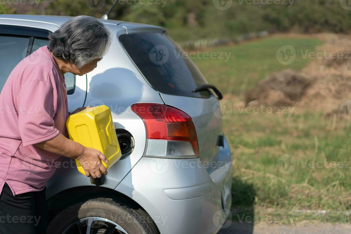 The car ran out of gas and stalled beside the road in suburbs and an elderly Asian woman used a gallon of spare gas to fuel the car. A woman prepares a gallon of spare gas to fuel before traveling. photo