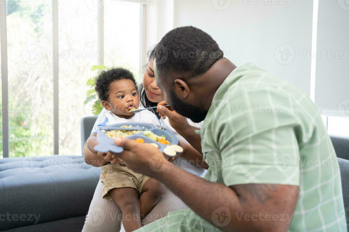 Asian mother feeding her 9 months old her cute little baby and African American helping for holding food plate At Home. Photo series of family, kids and happy people concept. Parents feed kids.