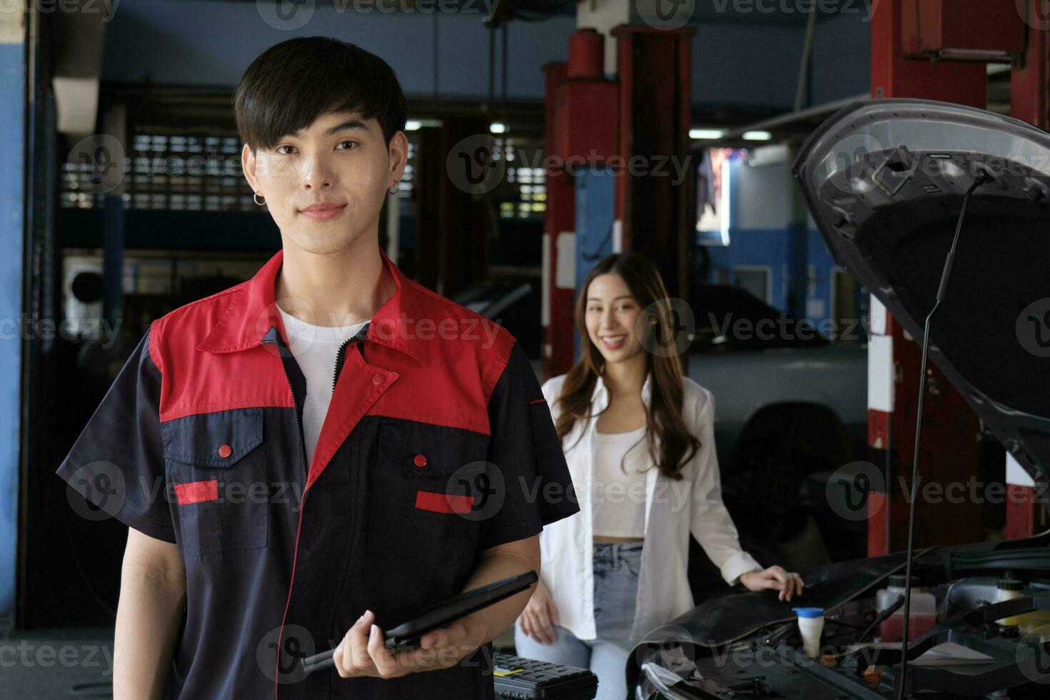 Portrait of young Asian male automotive mechanic worker with female customer's EV car at maintenance garage, happy smiles, professional service station, check and repair in auto transport industry. photo