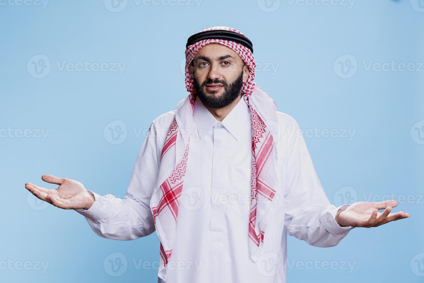 Confused muslim man wearing traditional clothes shrugging shoulders and looking at camera with uncertain expression. Arab person dressed in thobe spreading hands studio portrait photo