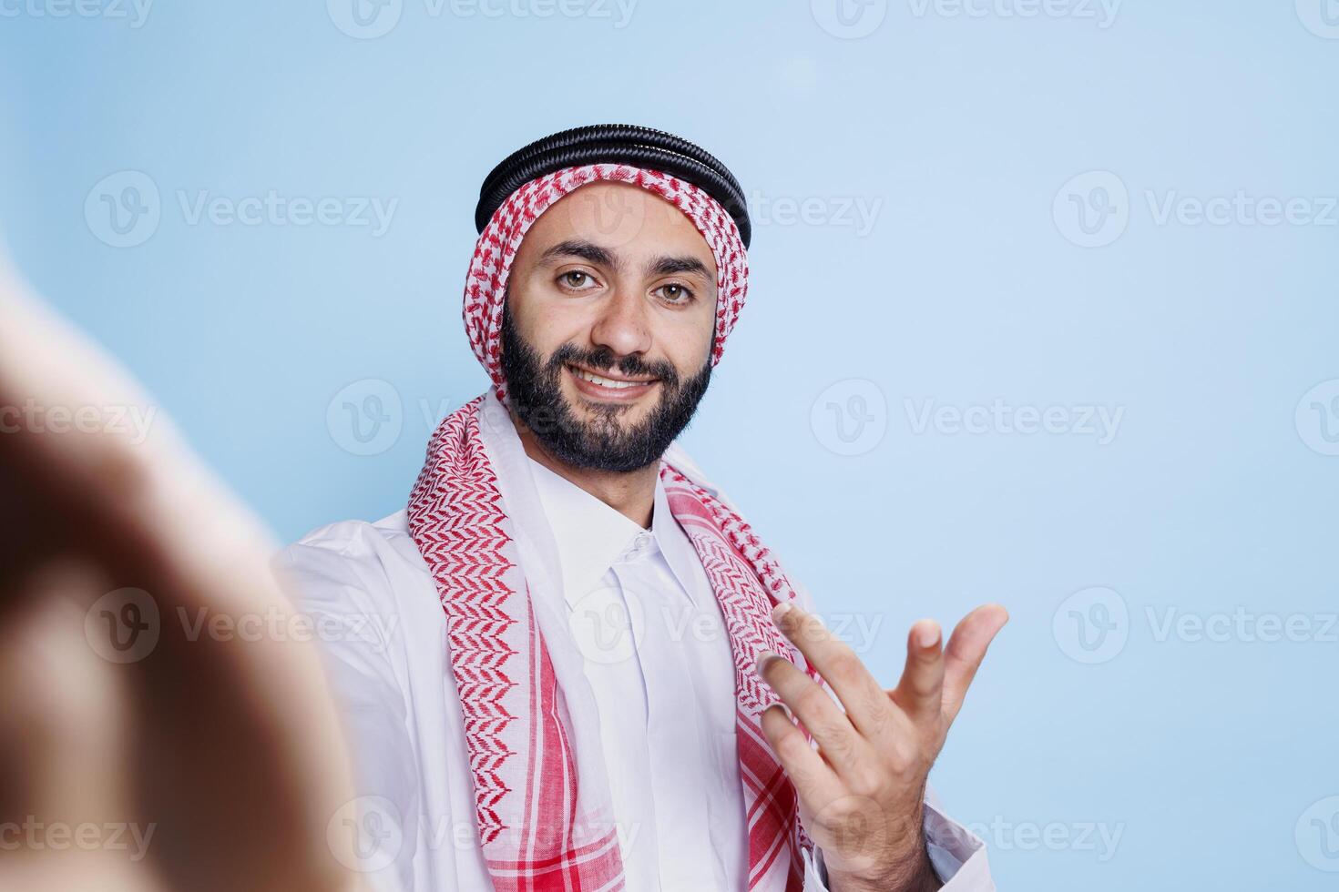 Happy smiling man dressed in traditional muslim clothes chatting during video conference fpv. Cheerful arab holding smartphone in hand and looking at camera first person view photo