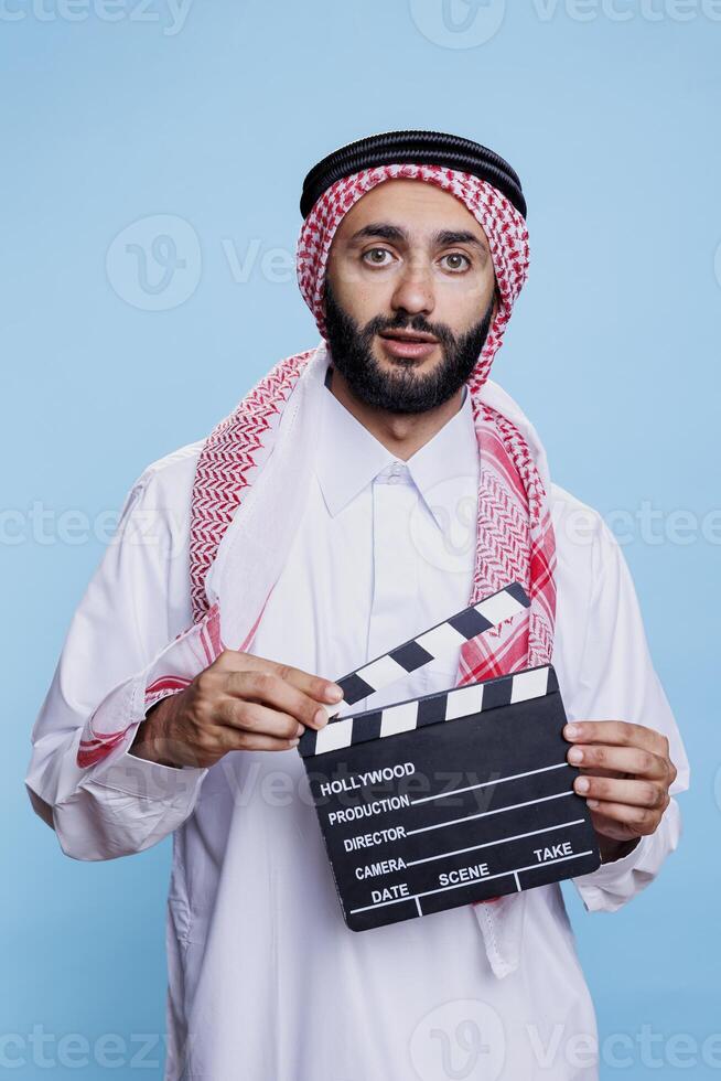 Movie scene director wearing traditional muslim clothes holding clapperboard and looking at camera. Arab dressed in islamic headscarf and thobe using clapboard studio portrait photo