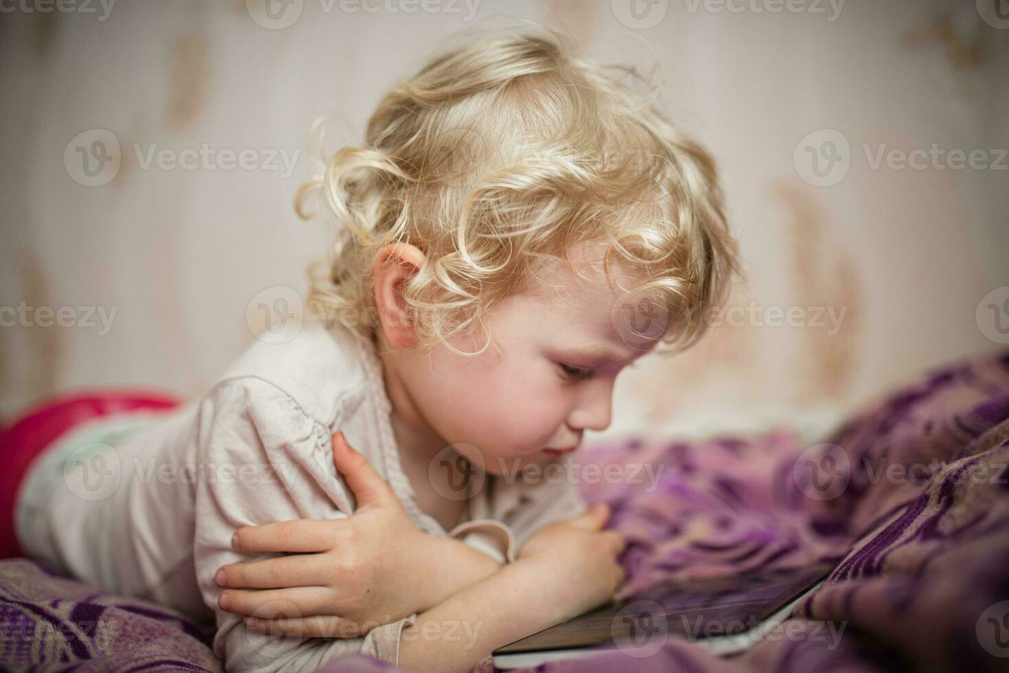 A little girl is lying on a couch with a tablet. The child watches cartoons instead of games and development on gadgets. A lonely child online photo