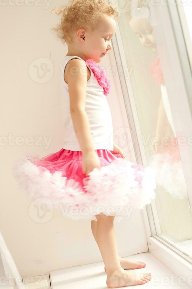 The little girl in a fluffy skirt standing at the open window. A child with white curly hair clings to the window handle. The little girl looks out the window. photo