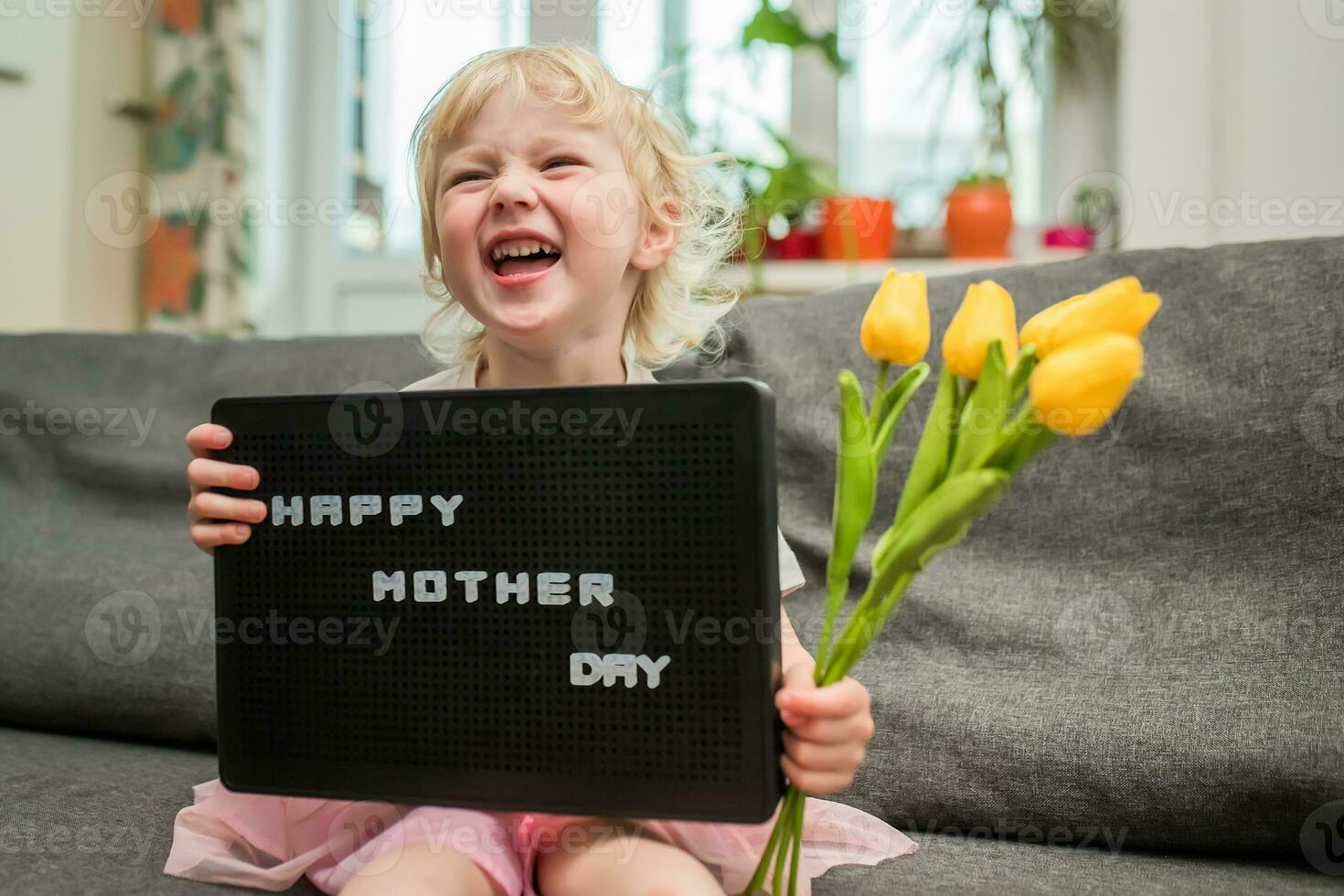 pequeño niña se ríe y sostiene un firmar con texto contento de la madre día. regalo para día festivo. hija sostiene un ramo de flores de amarillo tulipanes para mamá foto