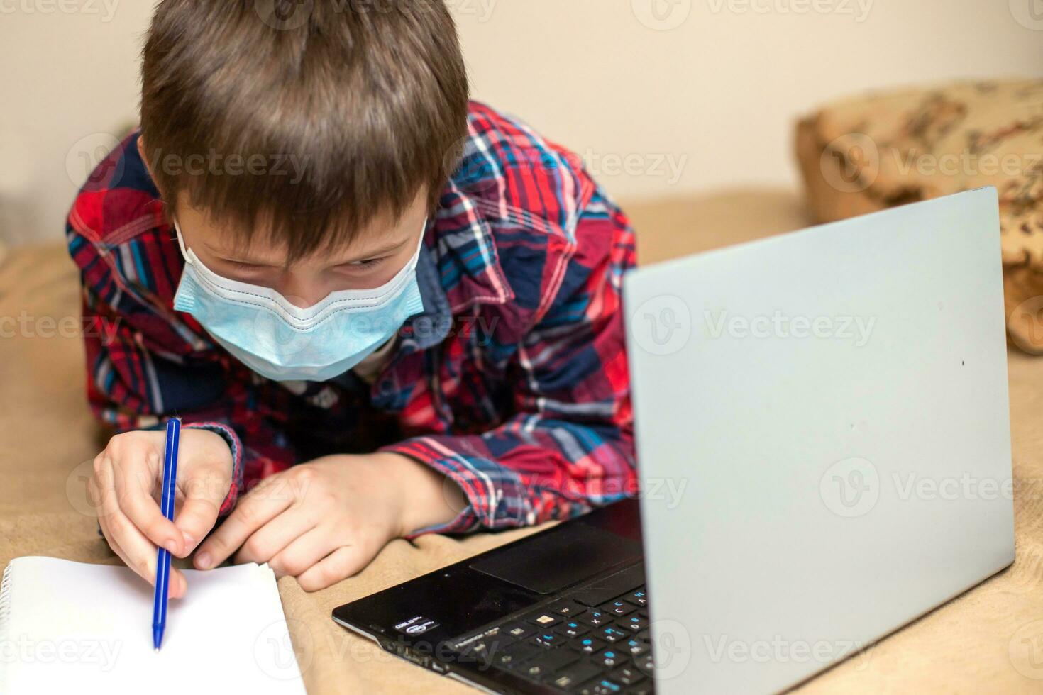 boy in protective medical mask lies near laptop and writes with left hand in notebook. child does lessons watching online broadcast lessons on Internet. e-learning, remote studying internet concept. photo