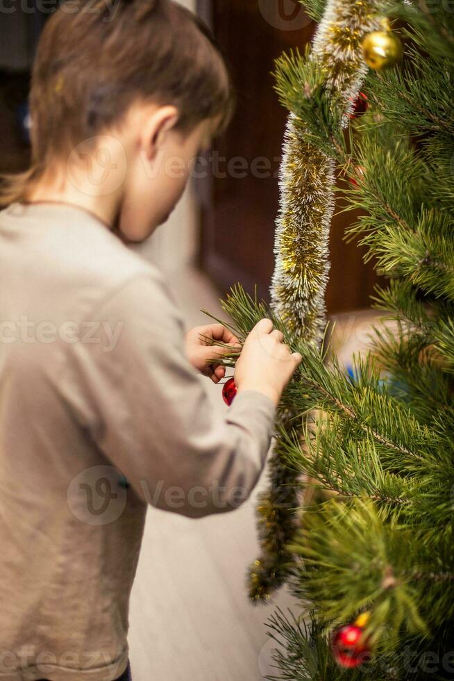 Boy clings a red ball on a branch of a Christmas tree at home. Preparing for Christmas at home. Real people. Decorating Christmas tree with a variety of toys photo