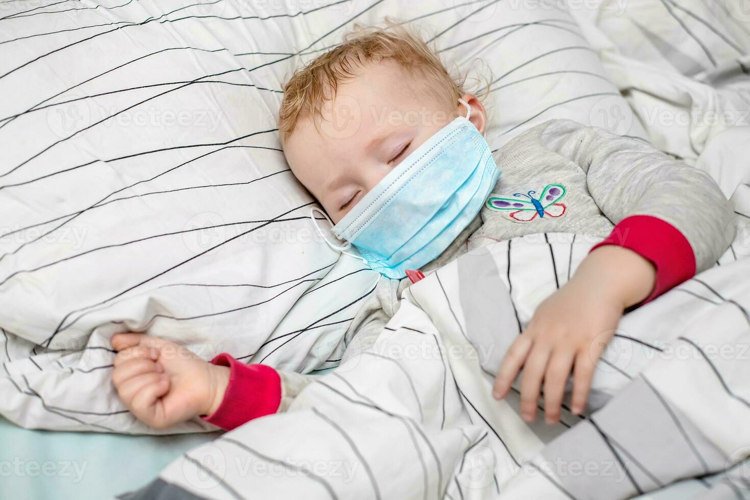 pequeño niño duerme en cama en protector médico mascarilla. enfermo niño en blanco cama. niño niñito uno años antiguo con gripe, influenza o frío protegido desde virus, contaminación entre pacientes con coronavirus foto