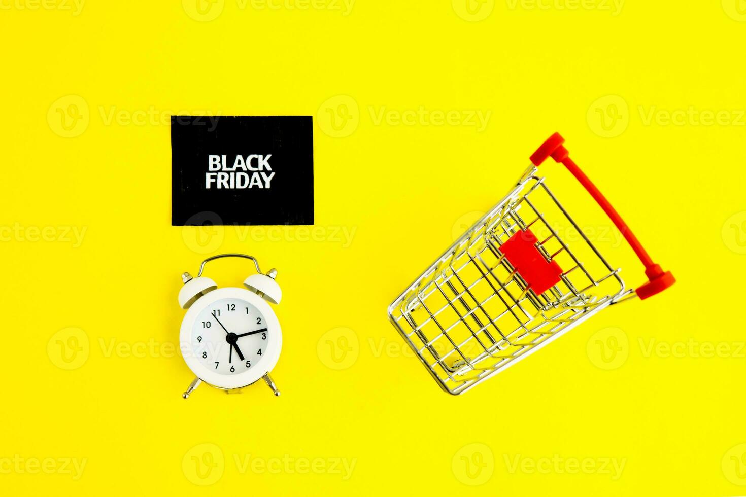 A white alarm clock, a shopping cart, and a black square that says black Friday on a yellow background. The topic of sales. photo