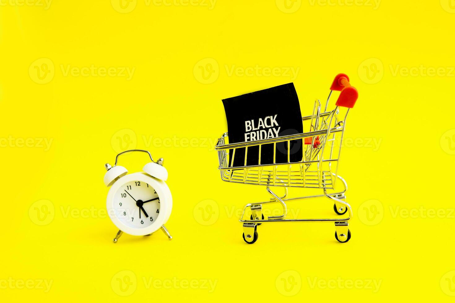 A white alarm clock, a shopping cart, and a black square that says black Friday on a yellow background. The topic of sales. photo