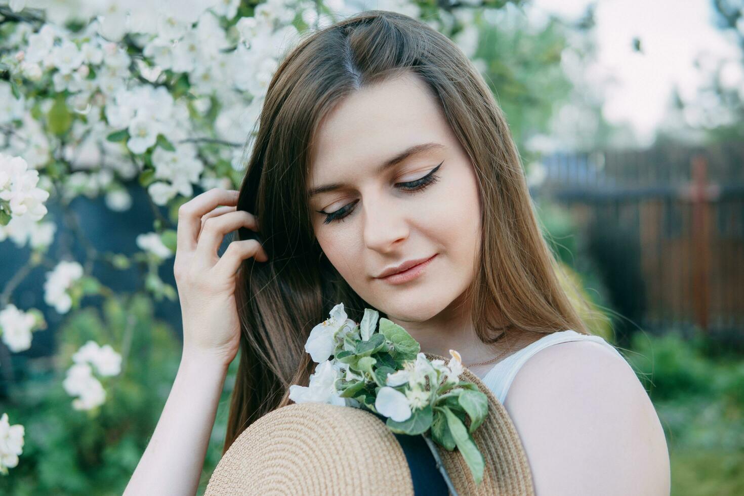 Beautiful young girl in white dress and hat in blooming Apple orchard. Blooming Apple trees with white flowers. photo